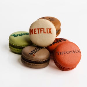 May include: A collection of colorful macarons, each with a different logo printed on top. The macarons are in shades of brown, green, orange, and beige. Logos include "Netflix", "Tropicana", "Escafe", "Tiffany & Co.", and a brown logo.