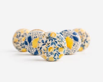 May include: A collection of five round ceramic knobs featuring a white base with a repeating pattern of yellow lemons and blue leaves. Each knob has a smooth, glossy finish and a circular shape, arranged in a cluster against a plain white background.