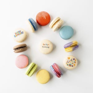 May include: A collection of colorful macarons arranged in a circle on a white surface. The macarons feature various colors, including blue, orange, pink, yellow, and brown. Some macarons have text that says "Happy Birthday" and "Emily's Birthday".