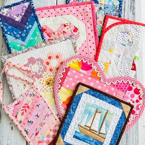 May include: A collection of colourful patchwork fabric pot holders, including a heart-shaped pot holder, a square pot holder with a sailboat design, and a rectangular pot holder with a floral design.
