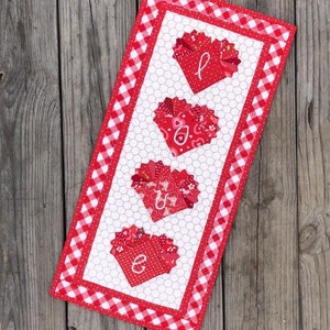 May include: A red and white gingham fabric wall hanging with a white background and four red hearts that spell out the word "LOVE".