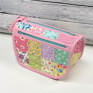 Pode incluir: Uma bolsa de cosméticos de tecido patchwork rosa com dois fechos de correr. A bolsa é feita de diferentes tecidos com padrões florais em tons de rosa, verde e azul.