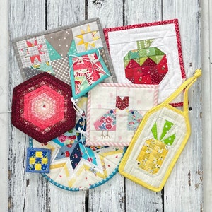 May include: A collection of colourful quilted pot holders featuring various designs, including a pineapple, a bird, an apple, and a hexagon. The pot holders are made with a variety of fabrics and patterns.