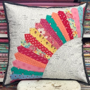 以下が含まれることがあります： 黒と白のドットの縁取りが付いた白い枕。枕の前面には、ピンク、赤、オレンジ、黄色、緑、青の色合いの布切れで作られたカラフルな扇形のデザインが施されています。布切れは、一番上の布切れが大きく、一番下の布切れが小さい、グラデーションパターンで配置されています。