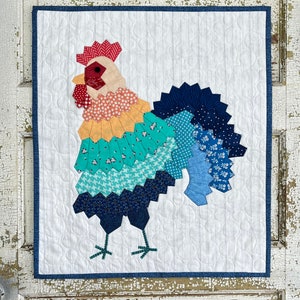Puede incluir: Una colcha de retazos colorida con un diseño de gallo. El gallo está hecho de varios tonos de azul, verde, amarillo, naranja y rojo. La colcha está enmarcada con un borde azul.