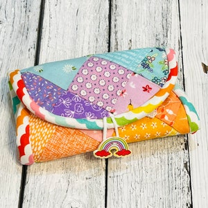 May include: A small, colorful patchwork fabric pouch with a rainbow charm. The pouch is folded closed and has a white cord with a tassel. The pouch is sitting on a white wooden surface.