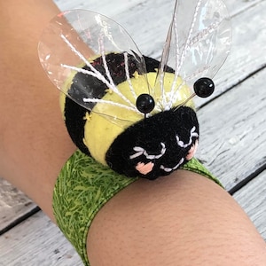 May include: A yellow and black bee with clear plastic wings is attached to a green fabric wristband. The bee has black eyes and a smiling face.