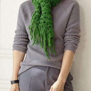 May include: A green crochet scarf with fringe detail is draped over a gray ribbed sweater. The scarf has an open, lacy design. The person is wearing gray pants and a black watch. The scarf adds a pop of color to the neutral outfit.
