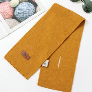 Puede incluir: Bufanda de color amarillo mostaza con forma rectangular y una peque&ntilde;a abertura. La bufanda tiene un dise&ntilde;o de punto texturizado y un parche de cuero marr&oacute;n con un logotipo. Gafas de sol y ovillos de lana en el fondo.