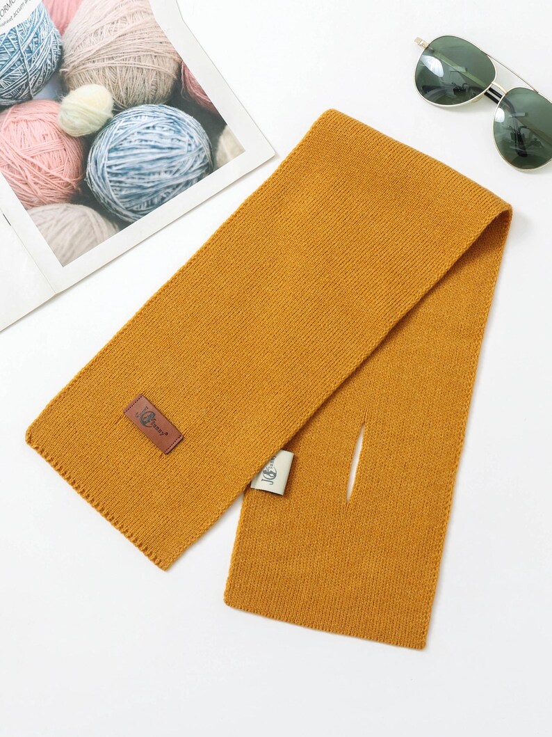 Puede incluir: Bufanda de color amarillo mostaza con forma rectangular y una peque&ntilde;a abertura. La bufanda tiene un dise&ntilde;o de punto texturizado y un parche de cuero marr&oacute;n con un logotipo. Gafas de sol y ovillos de lana en el fondo.