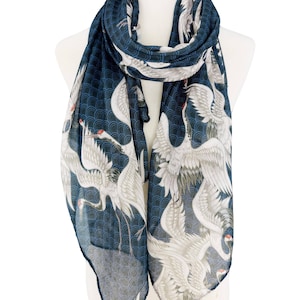 May include: A navy blue scarf featuring a repeating pattern of white cranes with red accents. The scarf is draped over a mannequin, showcasing the flowing fabric and detailed design. The background has a subtle wave pattern.