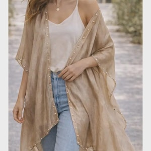 May include: A sheer, tan-colored kimono-style cover-up with pearl-like embellishments along the edges. The cover-up is worn over a white camisole and blue jeans. The garment has a flowing, lightweight appearance, suitable for layering.