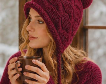 May include: A deep burgundy knit beanie featuring a textured cable pattern, a folded brim, and a matching pom-pom on top. The hat includes two braided side tassels with small pom-poms at the ends, crafted from a thick, warm yarn material.