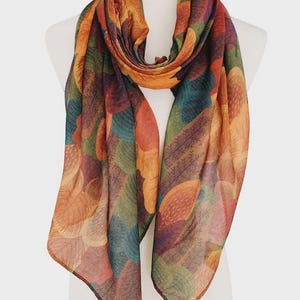 May include: A colorful scarf with a leaf pattern in shades of orange, green, blue, and purple. The scarf is draped over a white mannequin, showcasing its flowing design and vibrant colors. This accessory is perfect for adding a pop of color to any outfit.