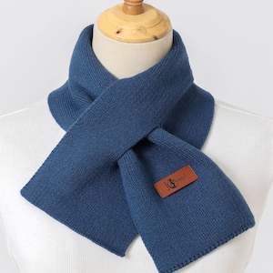 Puede incluir: Bufanda de punto azul marino con un parche rectangular de cuero con el texto "JG Sunny". La bufanda se muestra en un maniqu&iacute;, mostrando su dise&ntilde;o plegado. La bufanda est&aacute; hecha de un material suave y c&aacute;lido.