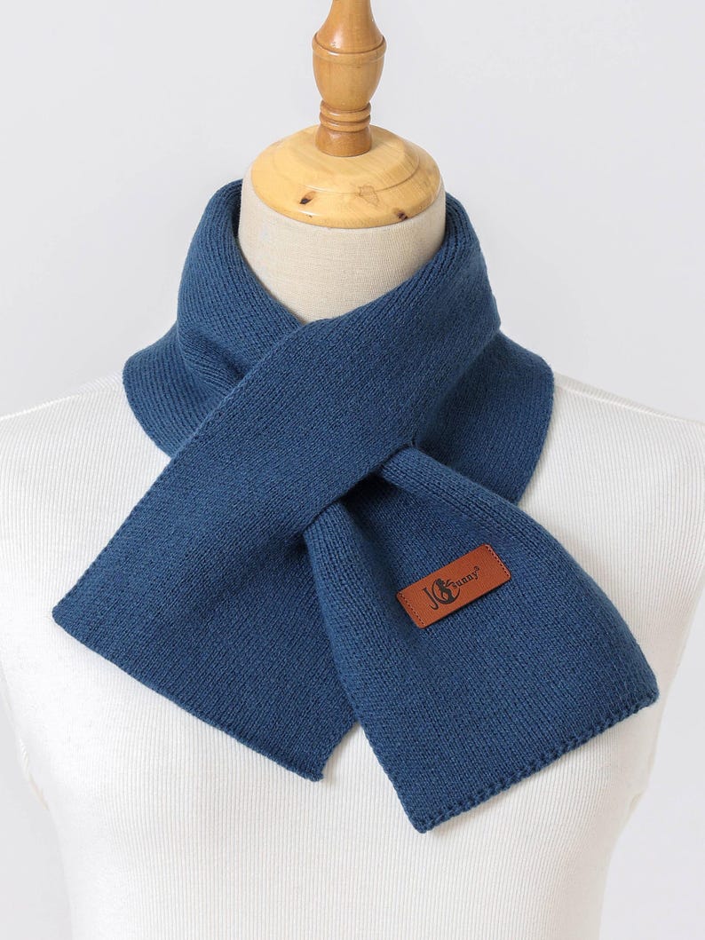 Puede incluir: Bufanda de punto azul marino con un parche rectangular de cuero con el texto "JG Sunny". La bufanda se muestra en un maniqu&iacute;, mostrando su dise&ntilde;o plegado. La bufanda est&aacute; hecha de un material suave y c&aacute;lido.