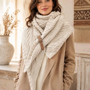 May include: A cream-colored triangular knit shawl featuring a textured cable pattern and a smooth brown leather strap with a metal buckle closure, worn over a tan coat and a white sweater.