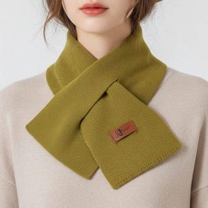 Puede incluir: Bufanda verde oliva con forma rectangular y parche de cuero. La bufanda es de punto y se lleva alrededor del cuello. La bufanda es de color liso.
