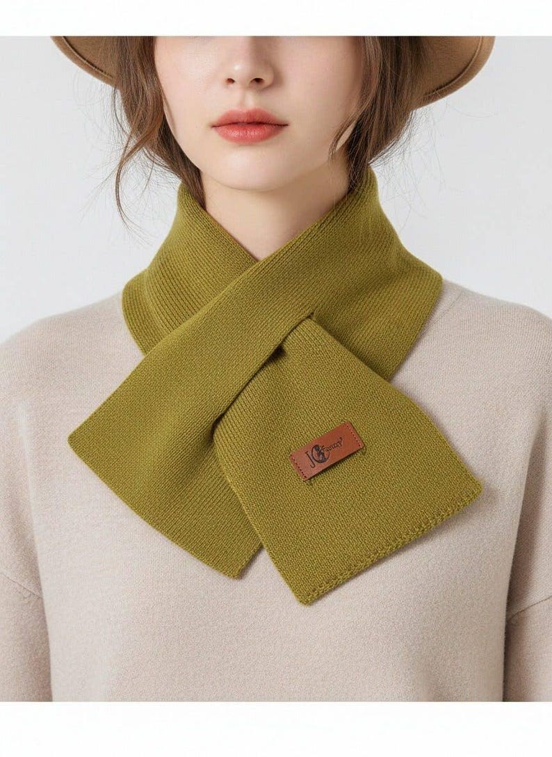 Puede incluir: Bufanda verde oliva con forma rectangular y parche de cuero. La bufanda es de punto y se lleva alrededor del cuello. La bufanda es de color liso.