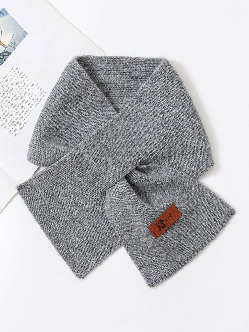 Puede incluir: Bufanda gris tejida con forma rectangular. La bufanda est&aacute; doblada sobre s&iacute; misma, creando un bucle. Una etiqueta de cuero marr&oacute;n con el texto "JG Sunny" est&aacute; adherida a la bufanda.