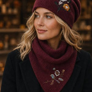 May include: A deep burgundy knit beanie and matching triangular scarf set. The beanie features a beaded floral embellishment with white and orange accents. The scarf displays a similar beaded floral design with blue, white, and brown tones.