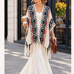 May include: An open-front poncho featuring a cream base with a geometric pattern in shades of red, teal, and dark blue. The garment has a wide, draped silhouette with long fringe detailing along the bottom hem, creating a textured appearance.