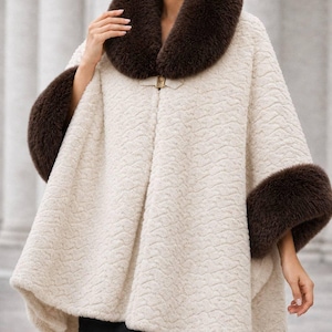 May include: A cream-colored textured cape featuring a dark brown faux fur collar and matching dark brown faux fur cuffs on the sleeves. The garment is secured at the neckline with a single gold-toned metal clasp closure, creating a structured silhouette.