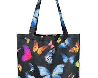 May include: Black tote bag featuring a vibrant butterfly pattern in shades of blue, orange, pink, and yellow. The bag has matching handles and a simple, rectangular shape, perfect for carrying everyday essentials.