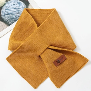 Puede incluir: Bufanda de punto amarillo mostaza con forma rectangular. La bufanda tiene un aspecto texturizado y una peque&ntilde;a etiqueta de cuero marr&oacute;n con el texto "JG Jenny". La bufanda est&aacute; doblada sobre s&iacute; misma.