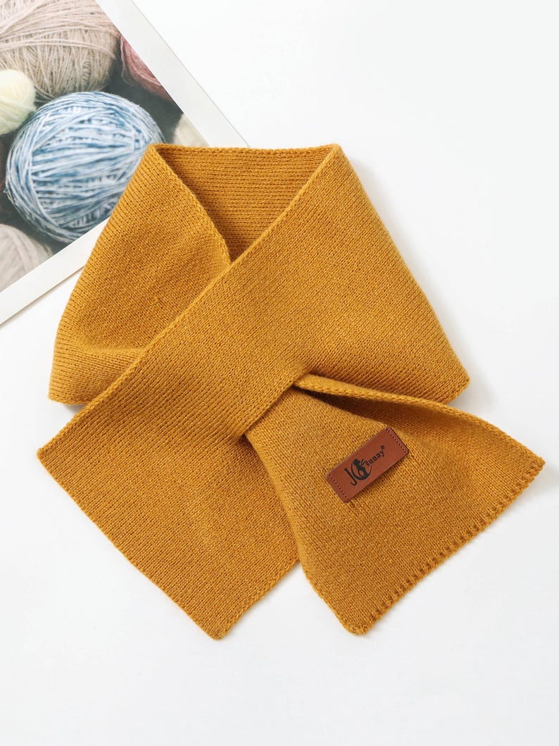 Puede incluir: Bufanda de punto amarillo mostaza con forma rectangular. La bufanda tiene un aspecto texturizado y una peque&ntilde;a etiqueta de cuero marr&oacute;n con el texto "JG Jenny". La bufanda est&aacute; doblada sobre s&iacute; misma.