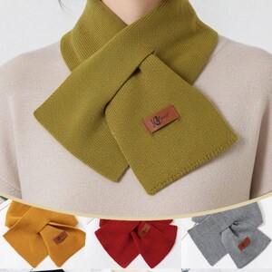 Puede incluir: Bufanda de punto verde oliva con dise&ntilde;o cruzado. La bufanda presenta un parche rectangular con un logotipo. Tambi&eacute;n se muestran bufandas adicionales en amarillo mostaza, rojo, gris, negro y crema.