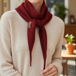 May include: A burgundy scarf with a subtle sparkle is tied around the neck. The scarf has long, pointed ends and a small, rectangular tag. The person is wearing a cream-colored sweater.