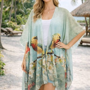 May include: A light blue kimono-style cover-up with a bird and floral print. The cover-up features a watercolor design with yellow, red, and white accents. The garment is worn over a white top and denim shorts. The background includes palm trees and a beach setting.