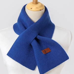 Puede incluir: Bufanda azul brillante de punto, exhibida en un maniqu&iacute;. La bufanda tiene una etiqueta de cuero marr&oacute;n con un logotipo. La bufanda est&aacute; doblada y drapeada, resaltando su textura y color. Un accesorio de invierno.