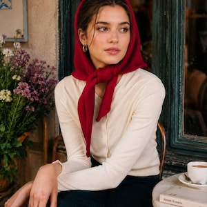 May include: A deep red knit headscarf tied under the chin, paired with a long-sleeved cream-colored button-down cardigan and dark blue denim jeans. The headscarf is made of a soft, textured fabric that frames the face, while the cardigan features a simple V-neckline and fitted sleeves.