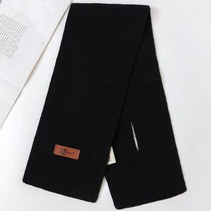 K&ouml;nnte beinhalten: Ein schwarzer Strickschal mit rechteckiger Form. Ein braunes Lederetikett mit dem Text "JG Teddy" ist auf den Schal gen&auml;ht. Der Schal ist zur H&auml;lfte gefaltet, mit einem Schlitz auf einer Seite.