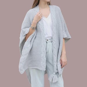 May include: A light gray knitted cape with a cable knit design on the sleeves and edges. The cape has a tassel detail and a draped, open front. The garment is made of a soft, textured material.
