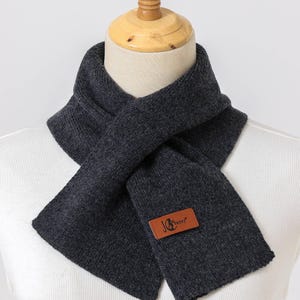 K&ouml;nnte beinhalten: Dunkelgrauer Schal aus Strickmaterial, auf einer Schaufensterpuppe pr&auml;sentiert. Der Schal ist um den Hals gewickelt und hat ein rechteckiges Etikett mit dem Text "JG Sunny" in Braun. Ein Winteraccessoire.