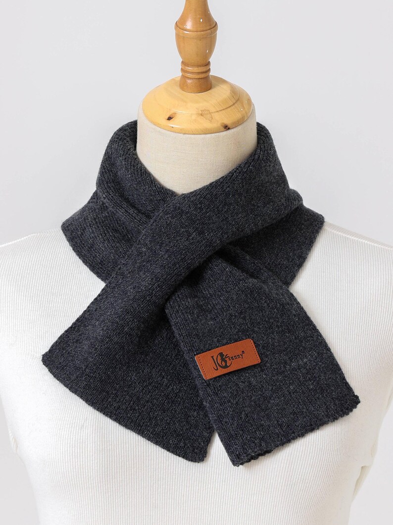 K&ouml;nnte beinhalten: Dunkelgrauer Schal aus Strickmaterial, auf einer Schaufensterpuppe pr&auml;sentiert. Der Schal ist um den Hals gewickelt und hat ein rechteckiges Etikett mit dem Text "JG Sunny" in Braun. Ein Winteraccessoire.