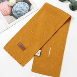 K&ouml;nnte beinhalten: Senfgelber Schal mit rechteckiger Form und kleiner &Ouml;ffnung. Der Schal hat ein strukturiertes Strickdesign und einen braunen Lederaufn&auml;her mit Logo. Sonnenbrille und Wollkn&auml;uel sind im Hintergrund.