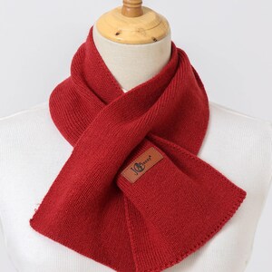 Puede incluir: Bufanda roja brillante de punto, exhibida en un maniqu&iacute;. La bufanda tiene un parche rectangular de cuero con un logotipo. Dise&ntilde;ada para llevar alrededor del cuello, ofrece calidez y estilo.