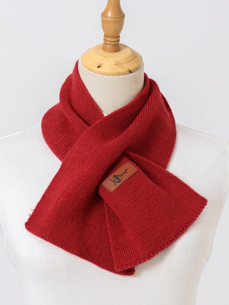 Puede incluir: Bufanda roja brillante de punto, exhibida en un maniqu&iacute;. La bufanda tiene un parche rectangular de cuero con un logotipo. Dise&ntilde;ada para llevar alrededor del cuello, ofrece calidez y estilo.