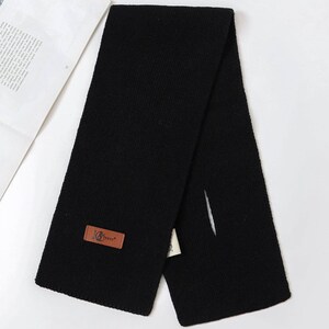 Puede incluir: Bufanda negra de punto con una etiqueta rectangular de cuero marr&oacute;n. La etiqueta tiene un logotipo y texto. La bufanda est&aacute; doblada por la mitad, con una ranura en un lado. Hecha de un material suave y c&aacute;lido.