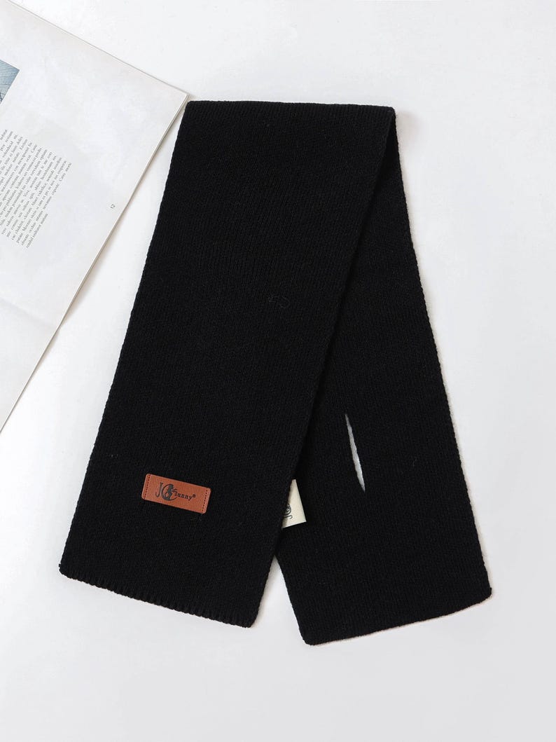 Puede incluir: Bufanda negra de punto con una etiqueta rectangular de cuero marr&oacute;n. La etiqueta tiene un logotipo y texto. La bufanda est&aacute; doblada por la mitad, con una ranura en un lado. Hecha de un material suave y c&aacute;lido.