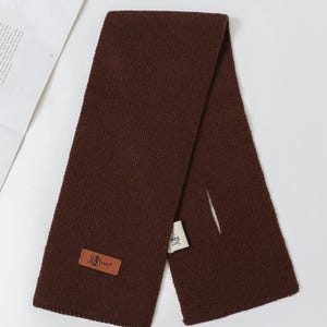 K&ouml;nnte beinhalten: Ein dunkelbrauner Strickschal mit einem rechteckigen Lederetikett mit der Aufschrift "JG Sunny". Der Schal ist gefaltet und liegt auf einer wei&szlig;en Oberfl&auml;che. Ein kleines wei&szlig;es Etikett ist am Schal befestigt.
