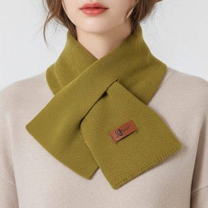 K&ouml;nnte beinhalten: Olivgr&uuml;ner Schal aus Strickstoff mit rechteckiger Form und Lederaufn&auml;her. Der Schal wird um den Hals getragen. Der Aufn&auml;her tr&auml;gt den Text "JG".