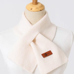 Puede incluir: Bufanda de color crema de punto, exhibida en un maniqu&iacute;. La bufanda tiene forma rectangular y una etiqueta de cuero marr&oacute;n con el texto "Jenny". La bufanda est&aacute; dise&ntilde;ada para llevarse alrededor del cuello.