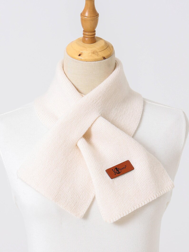 Puede incluir: Bufanda de color crema de punto, exhibida en un maniqu&iacute;. La bufanda tiene forma rectangular y una etiqueta de cuero marr&oacute;n con el texto "Jenny". La bufanda est&aacute; dise&ntilde;ada para llevarse alrededor del cuello.