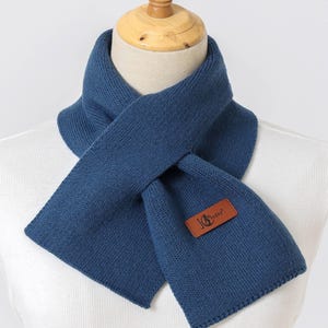 K&ouml;nnte beinhalten: Marineblauer Schal aus Strickstoff, pr&auml;sentiert auf einer Schaufensterpuppe. Der Schal hat ein rechteckiges braunes Lederetikett mit der Aufschrift "JG Sunny". Der Schal ist um den Hals der Schaufensterpuppe gelegt.