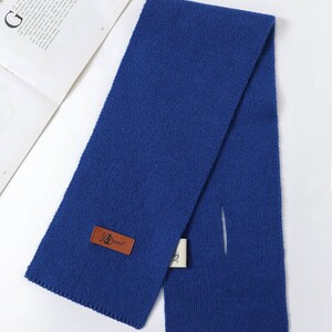 Puede incluir: Una bufanda de punto azul real con forma rectangular y una abertura de paso. Una etiqueta de cuero marr&oacute;n con el texto "JG Supply" est&aacute; adjunta. La bufanda est&aacute; doblada sobre una superficie blanca.